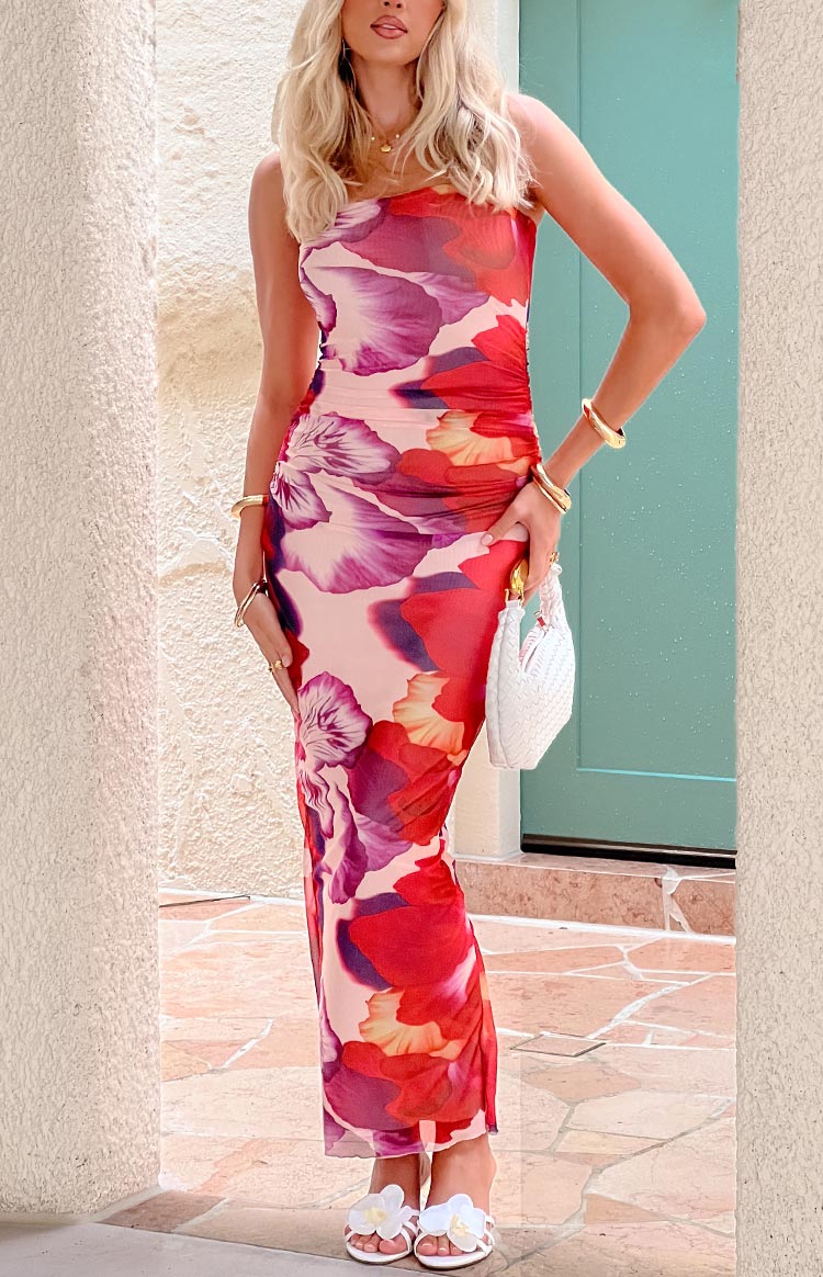 Model posing in a sunlit doorway- wearing a strapless maxi dress adorned with a bold purple- red- and pink floral print- featuring a ruched- figure-hugging bodice and a long- sheer mesh skirt.