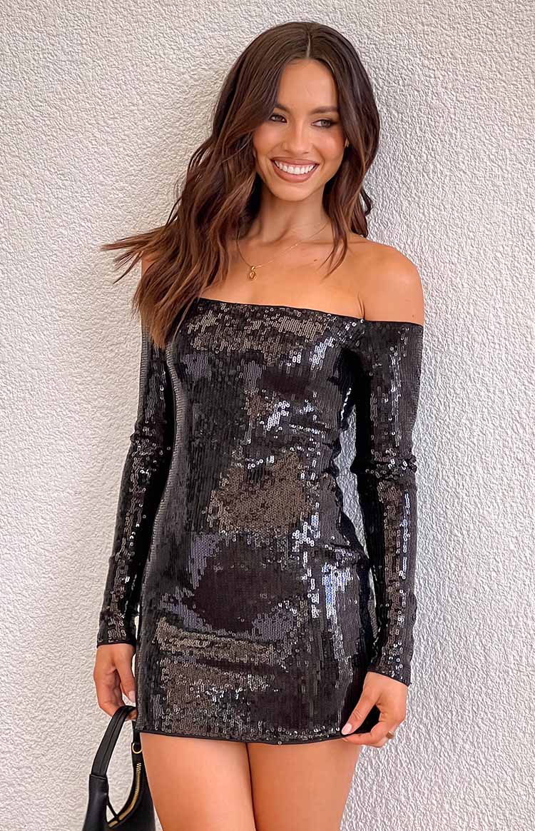 brunette girl wearing a black sequin long sleeve mini party dress with of shoulder neckline, styled with a gold necklace and black handbag
