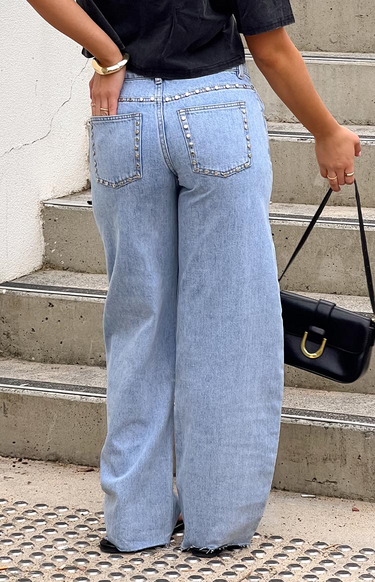 Back view of light wash denim low-rise jeans- featuring a wide-leg silhouette and silver studded detailing along the waist and pockets.