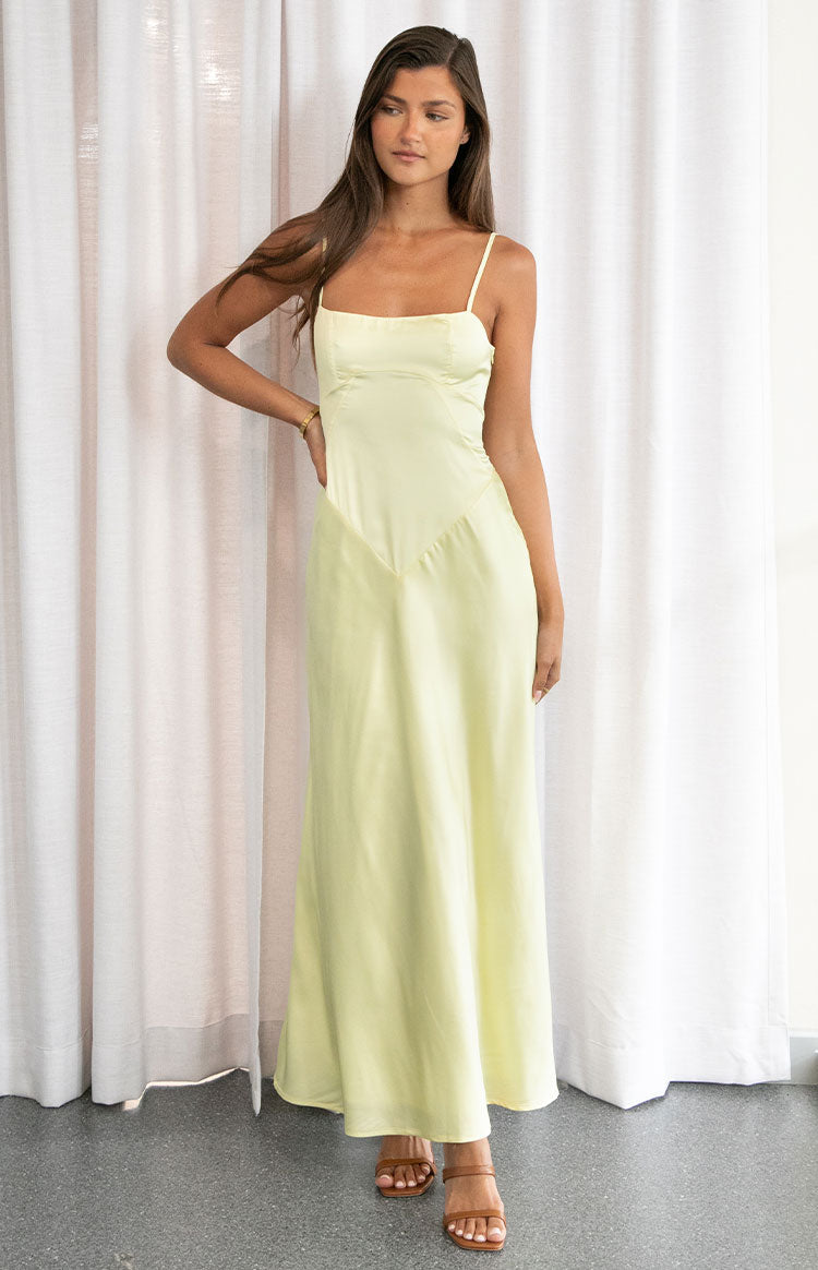 A female model wearing a pale yellow satin maxi dress with thin straps and a V-shaped seam at the waist.