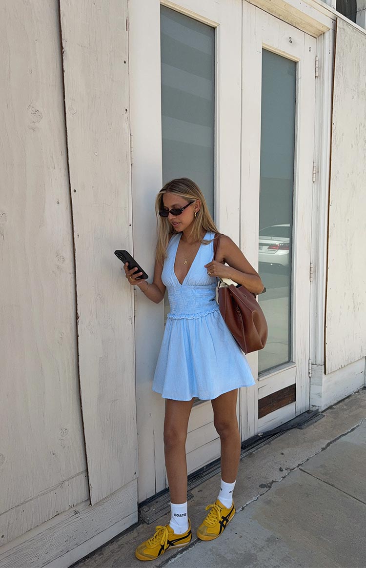 A woman wears a sleeveless mini dress in a light blue and white pinstripe fabric. The dress is designed with a V-neck- a shirred waist- and an A-line skirt- and is paired with white socks and bright yellow sneakers.
