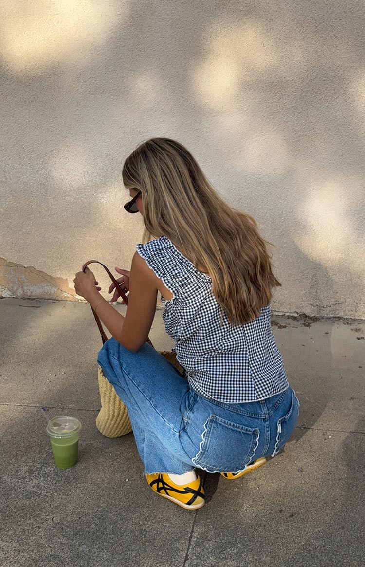 A woman crouches to show the back detail of her mid-wash jeans- which feature a white scalloped embroidered trim along the top of the back pockets. She is wearing a blue and white gingham top and yellow sneakers.