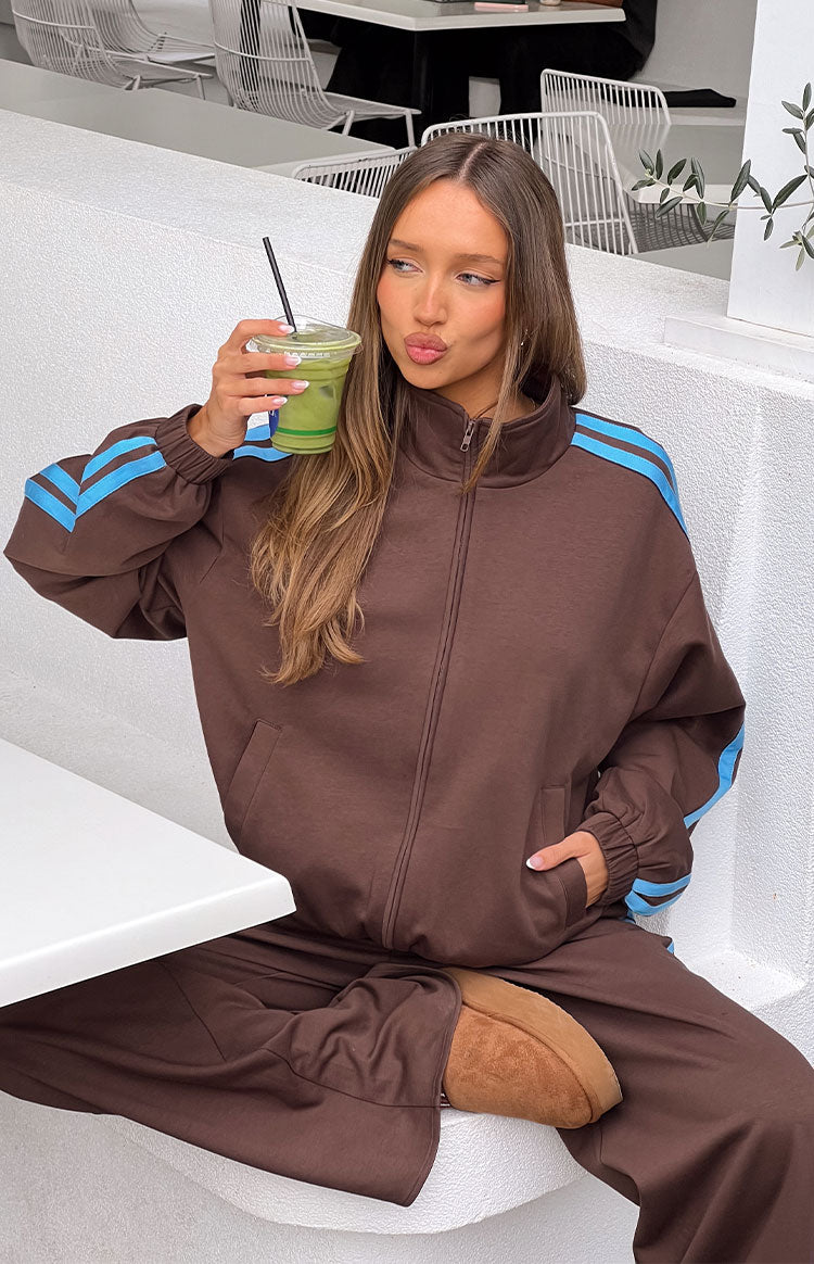 A female model wearing an oversized brown zip-up track jacket with a high collar and light blue stripes down the arms.