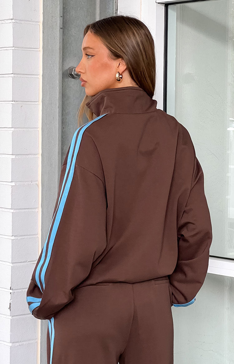 A back view of our model wearing an oversized brown zip-up track jacket with a high collar and light blue stripes down the arms. Perfectly wear with a matching pants.