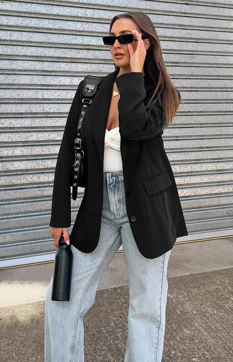 Brunette model wearing a black oversized blazer. The blazer has immitation pockets on the front with 1 large button on front seam for accessible closure.