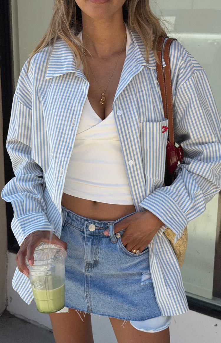 Model wearing an oversized blue and white striped button-up shirt- styled open over a white top and a denim mini skirt. The shirt has a small red embroidered detail on the chest pocket.