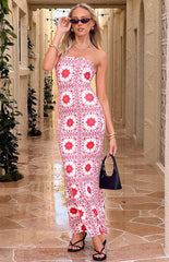 A person with blonde hair and sunglasses models a white strapless maxi dress with a red geometric pattern. They are wearing gold jewelry and carrying a small black handbag while standing in a sunlit outdoor corridor.