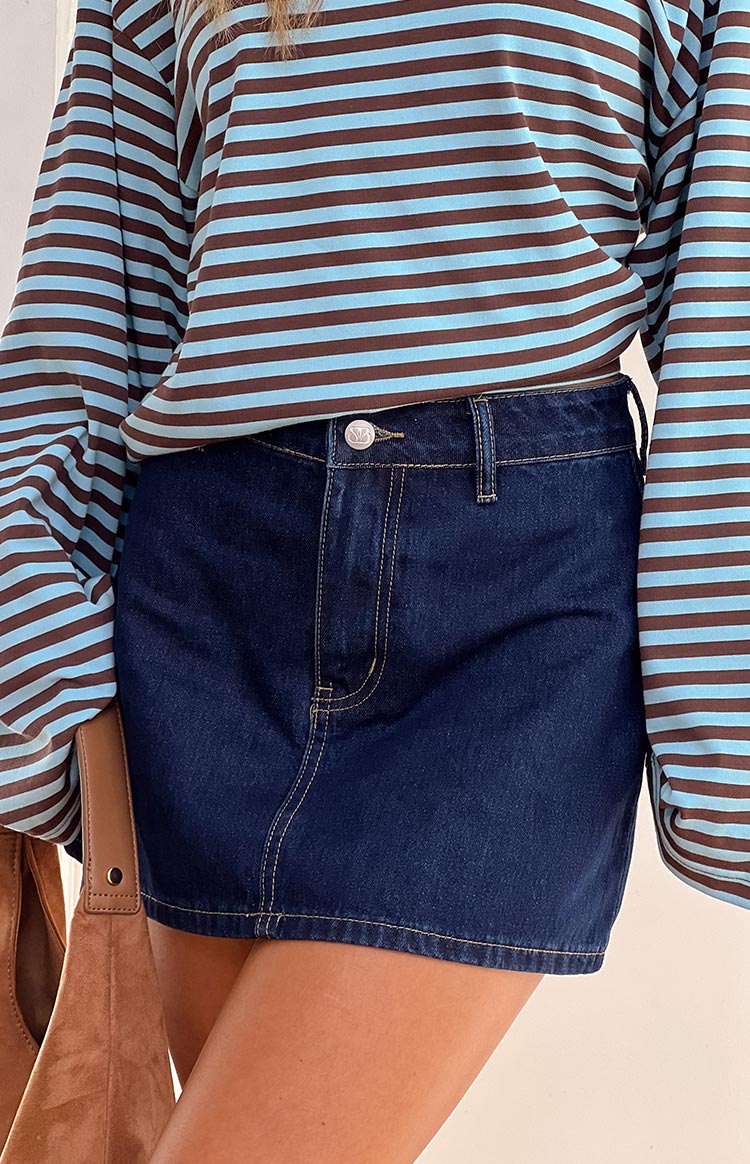 A close-up shows a person wearing a blue and brown horizontally striped long-sleeved shirt paired with a dark blue denim mini skirt. A brown shoulder bag is partially visible on the left.
