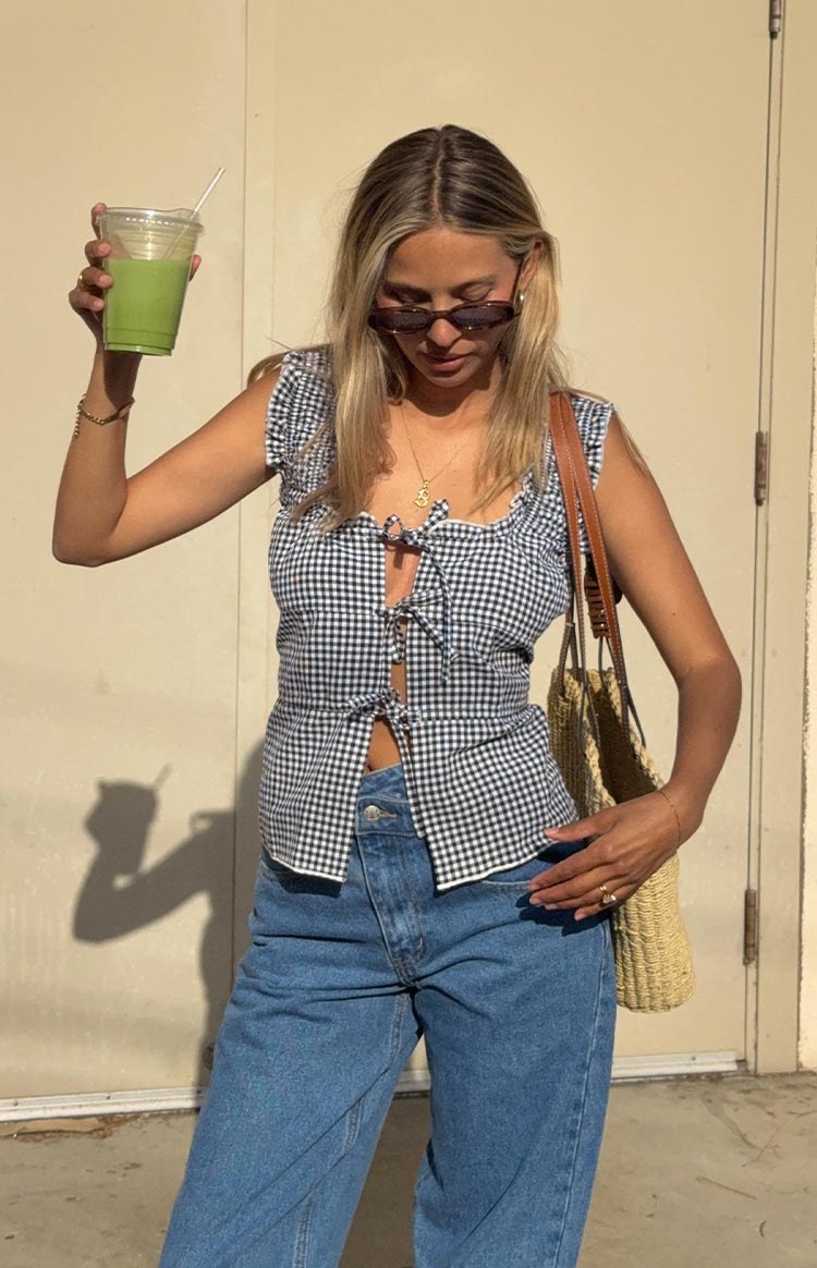 Model wearing a black and white gingham top with ruffled cap sleeves. The top has two tie-front details creating keyhole cutouts and is paired with blue jeans.