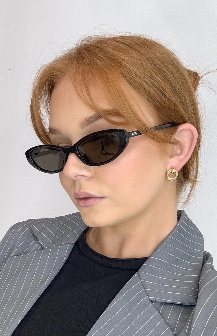 Black thin cat-eye sunglasses with dark lenses- paired with a grey pinstripe blazer and a black top.