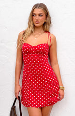 Model wearing a vibrant red mini dress with white polka dots- a sweetheart neckline- and thin tie-straps- accessorized with gold jewelry and a dark bag.
