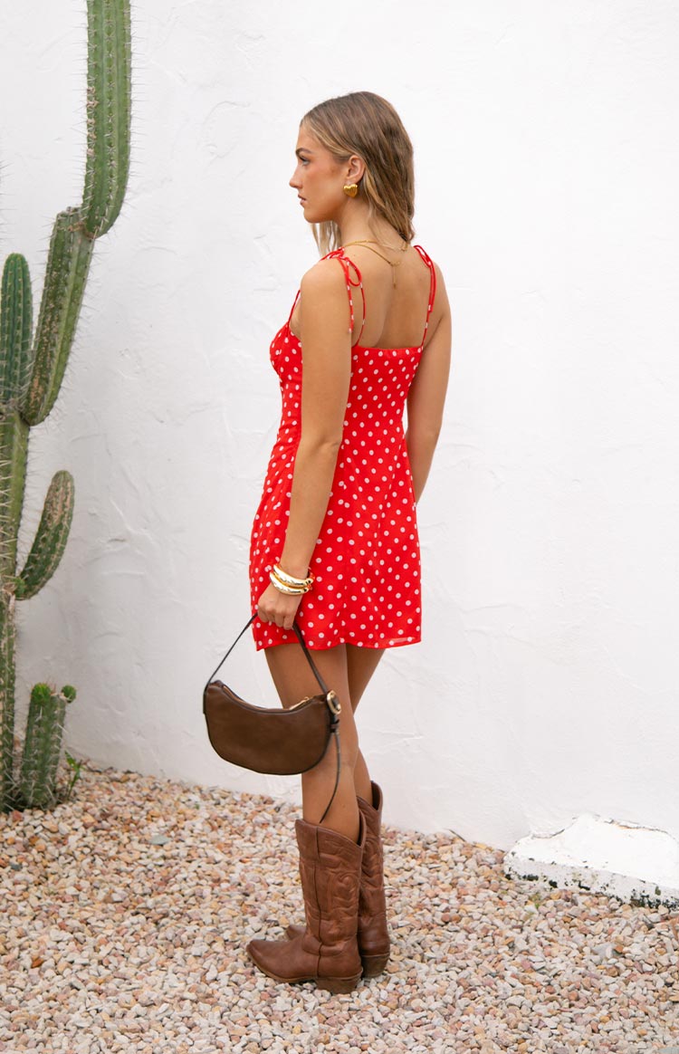 Model wearing a vibrant red mini dress with white polka dots- a sweetheart neckline- and thin tie-straps- accessorized with gold jewelry and a dark bag.