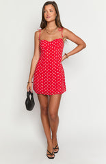 Model wearing a vibrant red mini dress with white polka dots- a sweetheart neckline- and thin tie-straps- accessorized with gold jewelry and a dark bag.