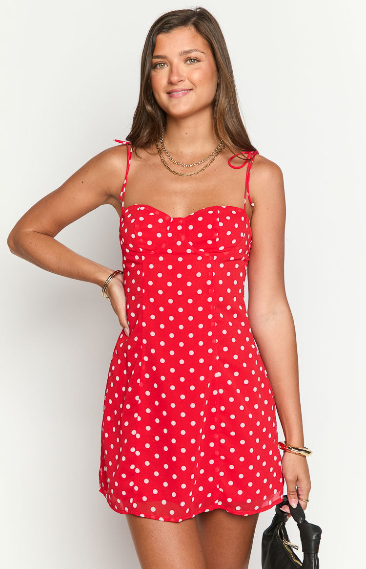 Model wearing a vibrant red mini dress with white polka dots- a sweetheart neckline- and thin tie-straps- accessorized with gold jewelry and a dark bag.