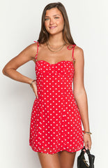 Model wearing a vibrant red mini dress with white polka dots- a sweetheart neckline- and thin tie-straps- accessorized with gold jewelry and a dark bag.