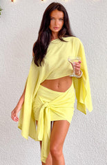 Brunette model wearing a yellow one shoulder flowy mini dress with a tie wrap skirt and holding a drink.