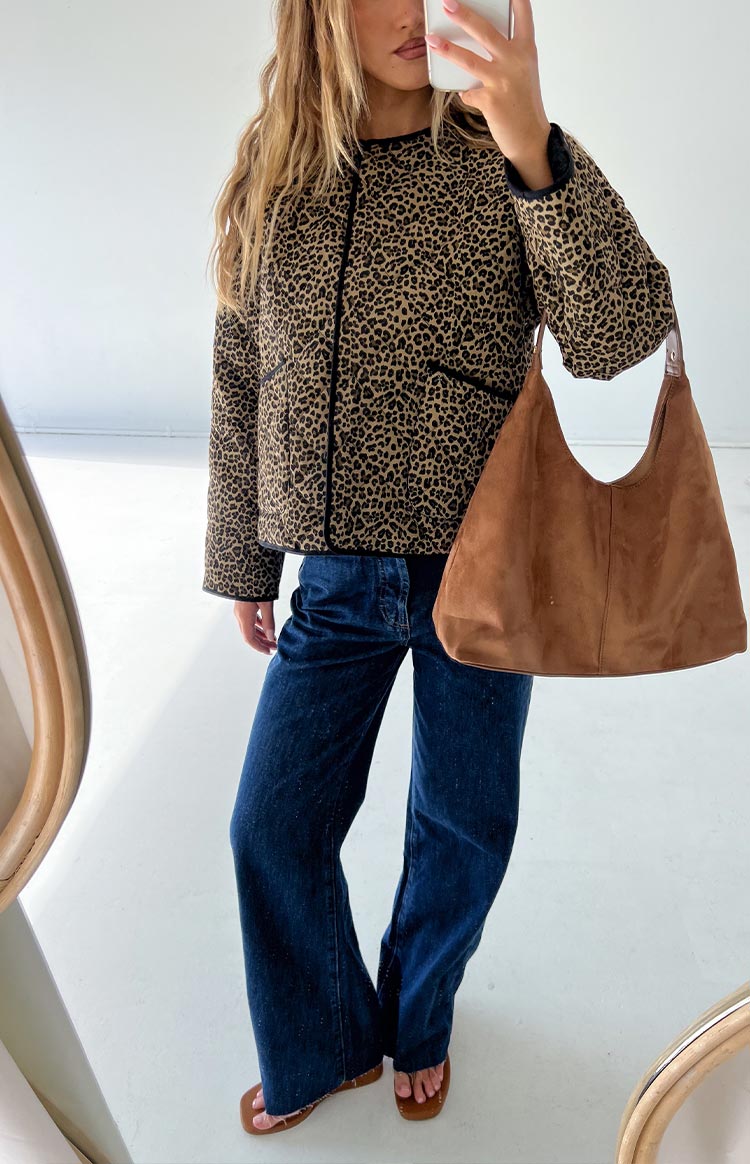 Model wearing a leopard print quilted jacket with black trim- dark blue wide-leg jeans- and brown sandals- holding a large brown slouch bag.
