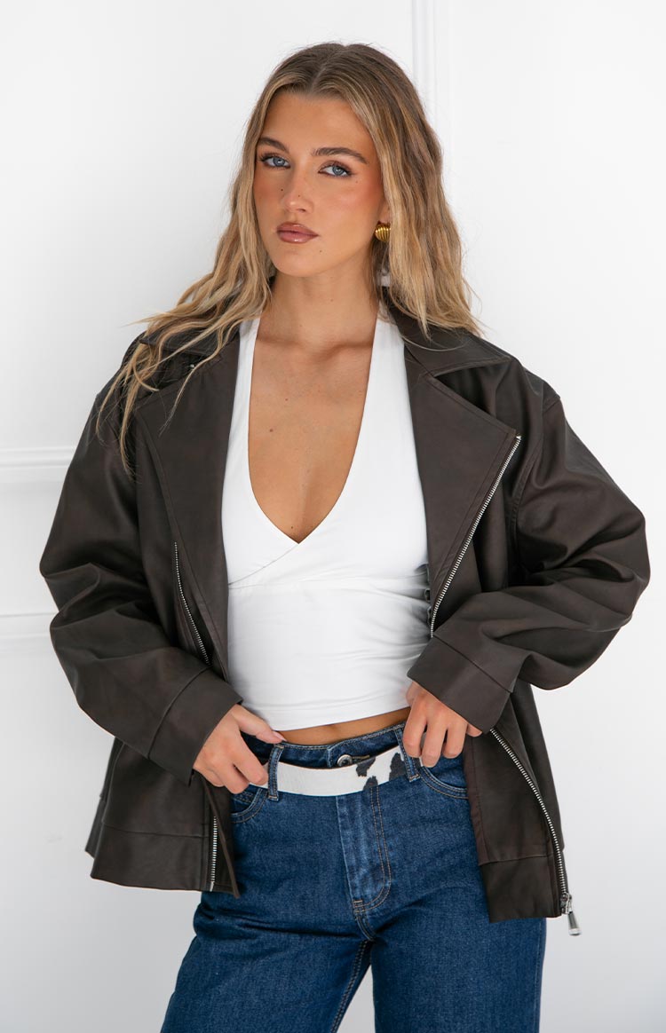 Model wearing an open dark grey or black oversized leather-look jacket- a white halter crop top- and blue jeans with a white belt.