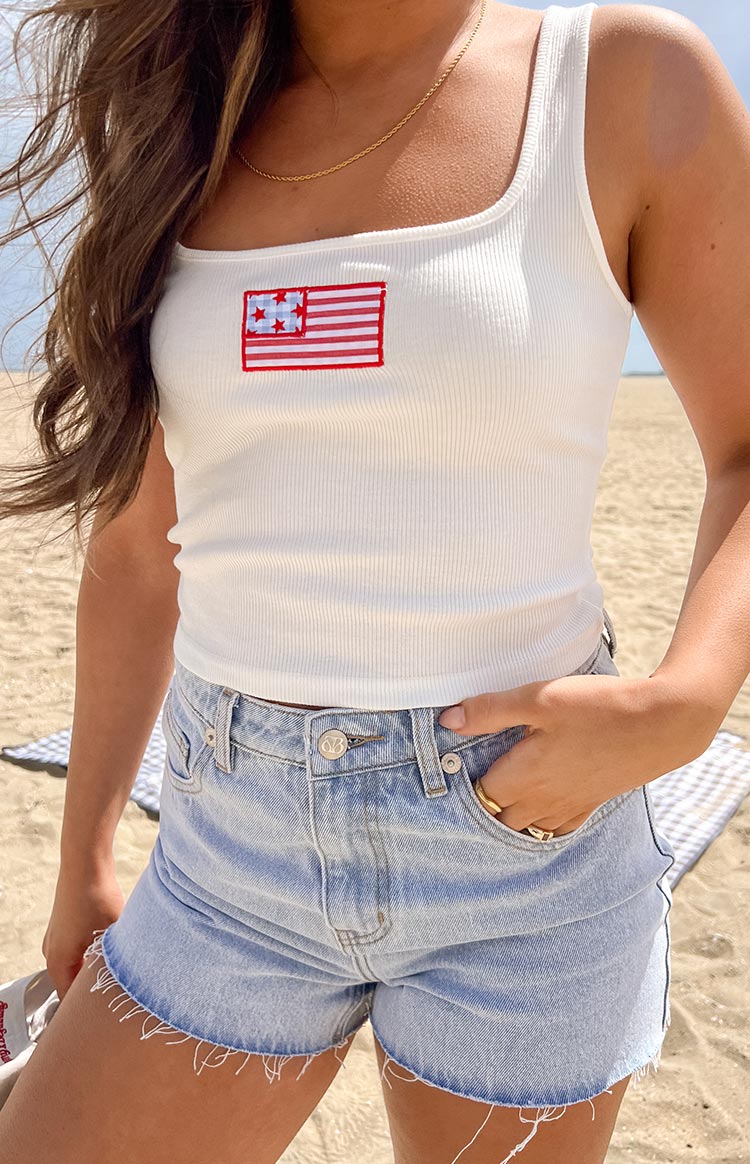 Girl wearing a white tank top with a USA flag embroidered patch paired with light wash denim shorts