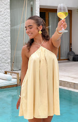 Girl is smiling and raising her glass to the camera. She is wearing a yellow mini dress and gold seashell earrings.