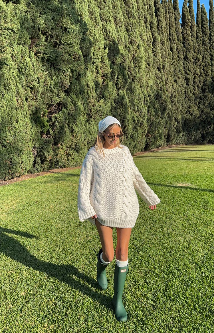 Model walking in a park wearing an oversized cream cable-knit sweater as a mini dress. The chunky knit sweater has a crew neck and is paired with green rain boots.