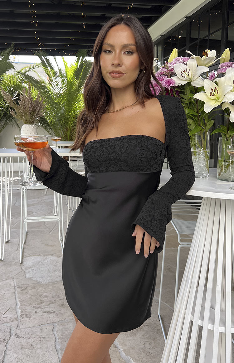 Brunette model wearing black long sleeve mini dress and holding a drink in front of white tables.