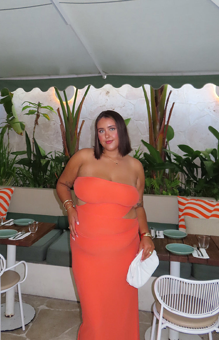 Brunette girl is wearing an orange maxi dress and gold starfish earrings. SHe is holding a drink and wearing white flip flop sandals.