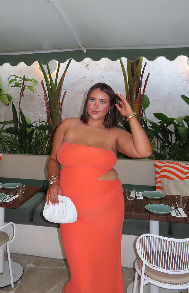 Brunette girl is wearing an orange maxi dress and gold starfish earrings. SHe is holding a drink and wearing white flip flop sandals.