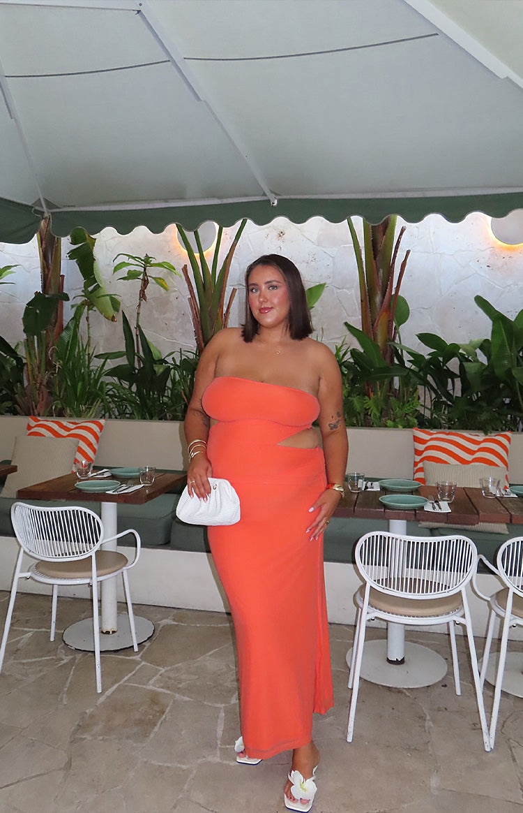 Brunette girl is wearing an orange maxi dress and gold starfish earrings. SHe is holding a drink and wearing white flip flop sandals.